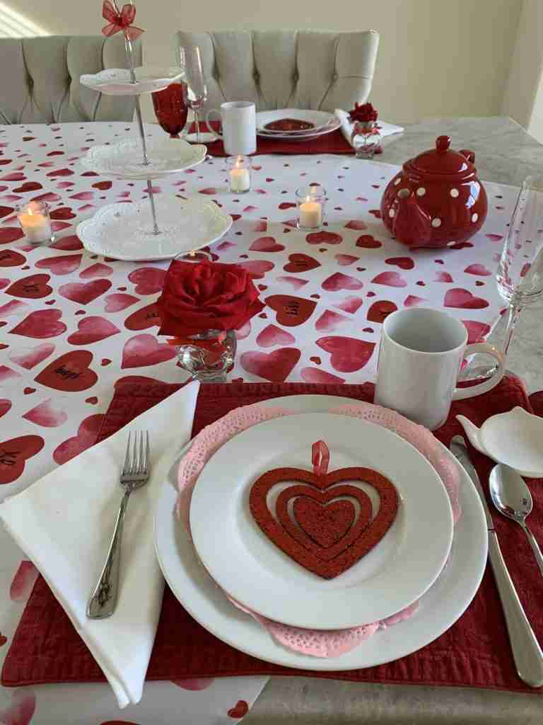 A Valentine's Day tablescape with a pretty heart-shaped tablecloth, white plates, pink doilies, red placemats, little vase polka dot teapot.