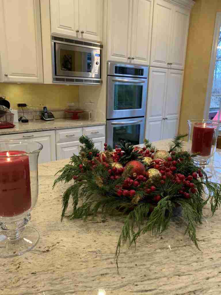 Red pillar candles in glass hurricanes, greenery in a wooden bowl with ornaments, and red berries.