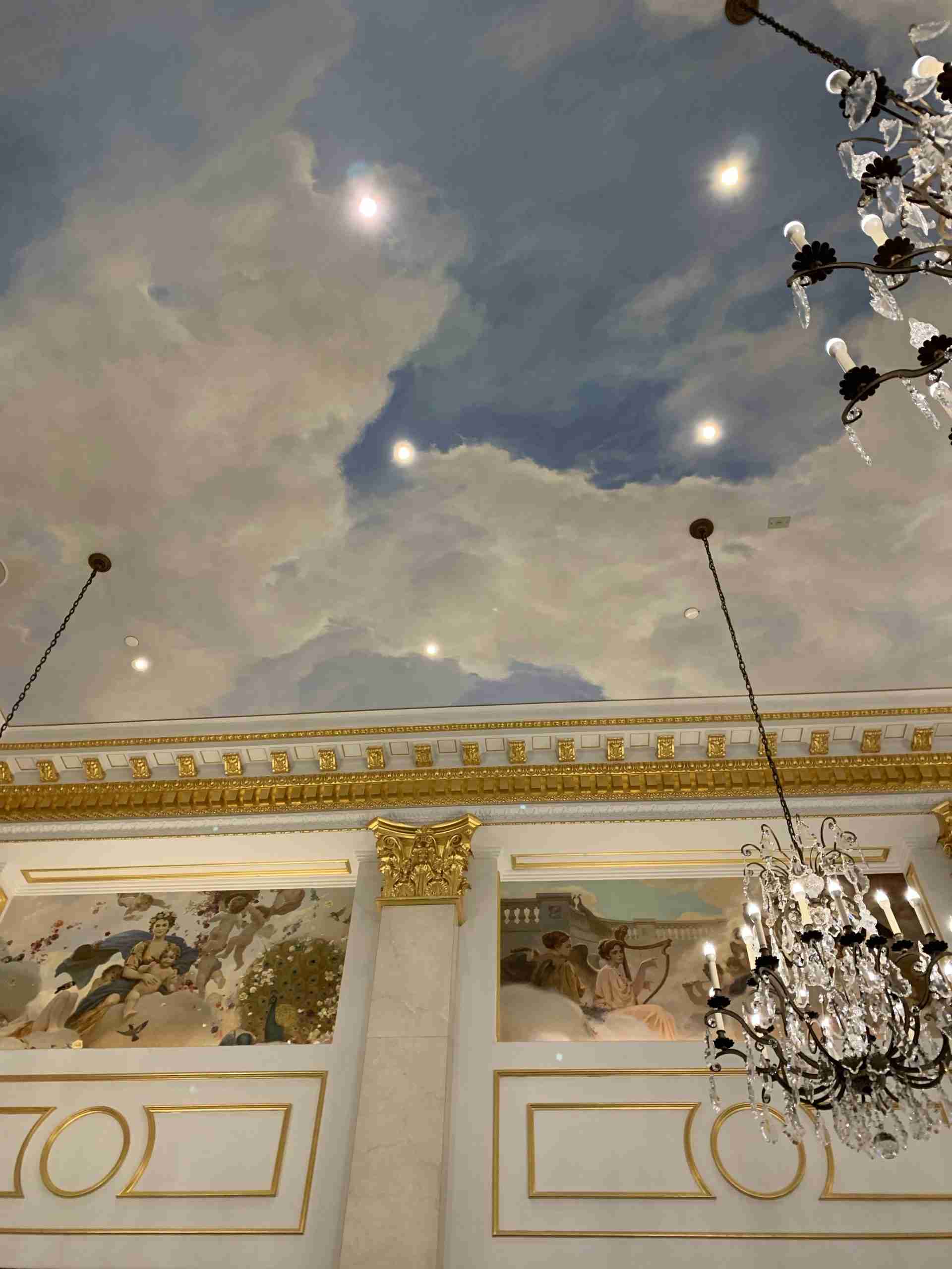 a mural of blue sky with clouds and a crystal chandelier at Astor Court in the St. Regis hotel.