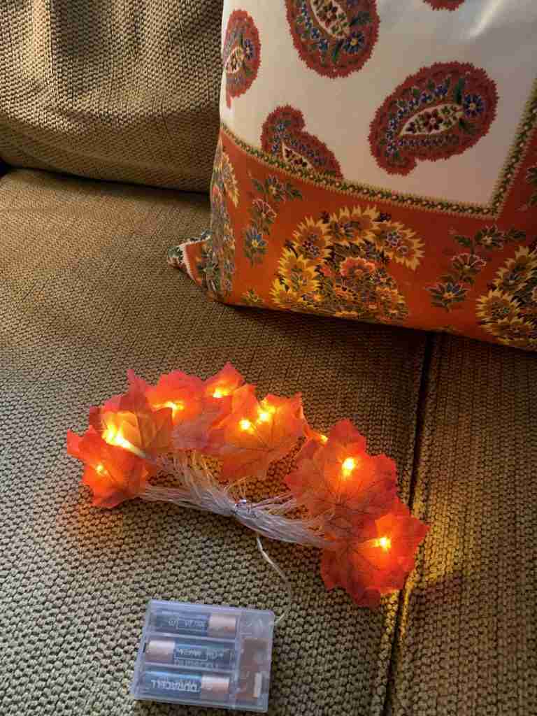 A photo of the leaf lights which are a strand of battery-operated white lights with silk leaves glues to the lights.  In the image, there is a beautiful autumnal paisley throw pillow on a neutral chenille sofa.