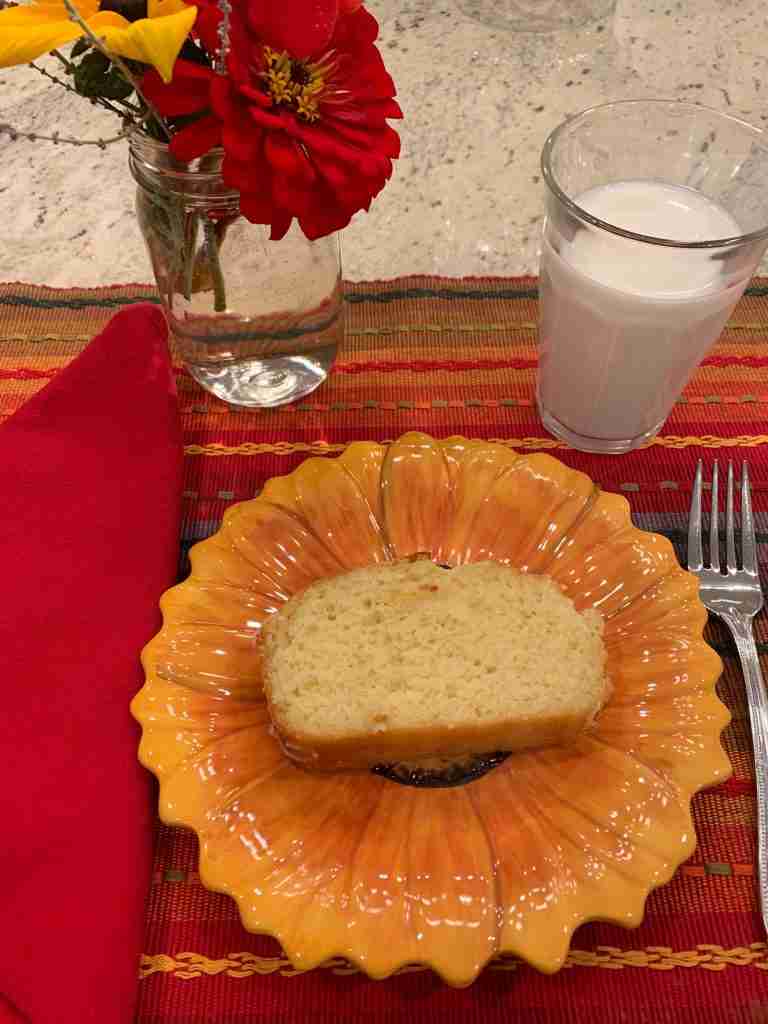 A bright striped placemat with a glass of milk and a piece of lemon cake on a sunflower-shaped cake plate.  There is also a mason jar of zinnias.