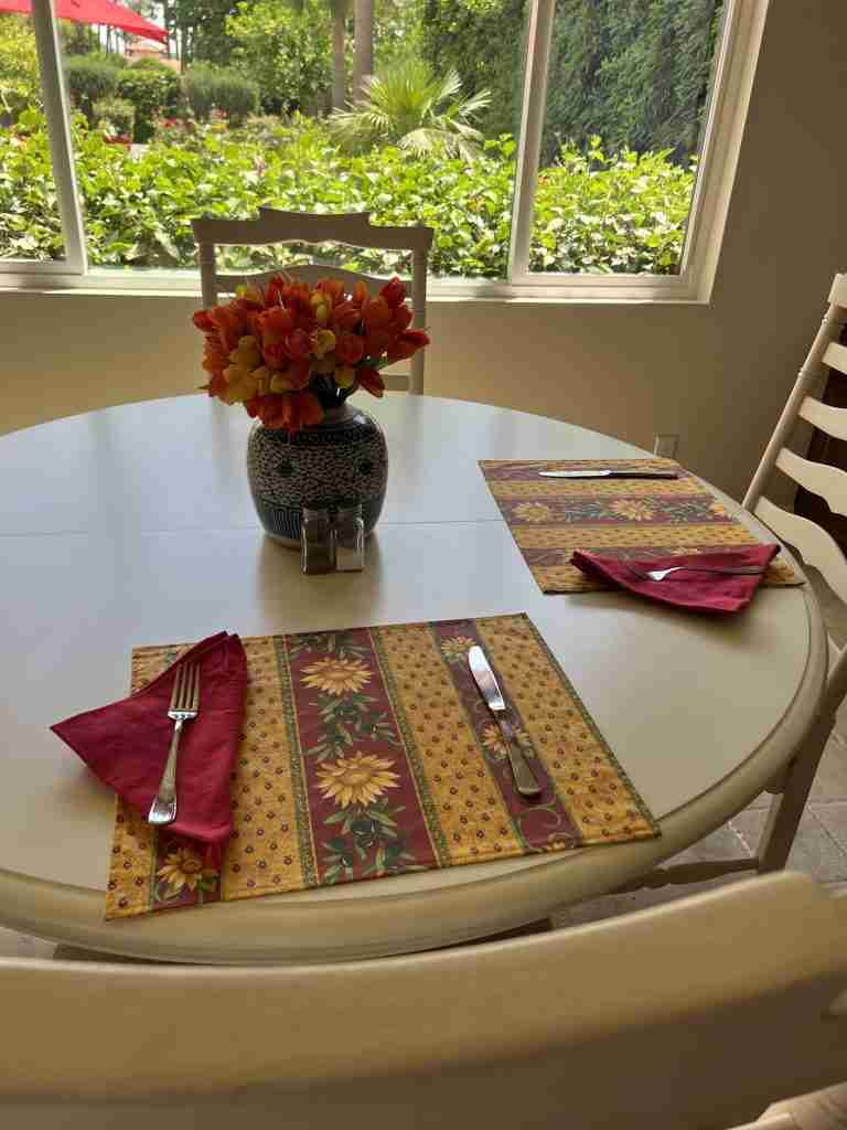 A white pedestal table with white ladderback chairs, sunflower placemats, red napkins and a blue and white ginger jar on the table containing red, yellow, and orange tulips.