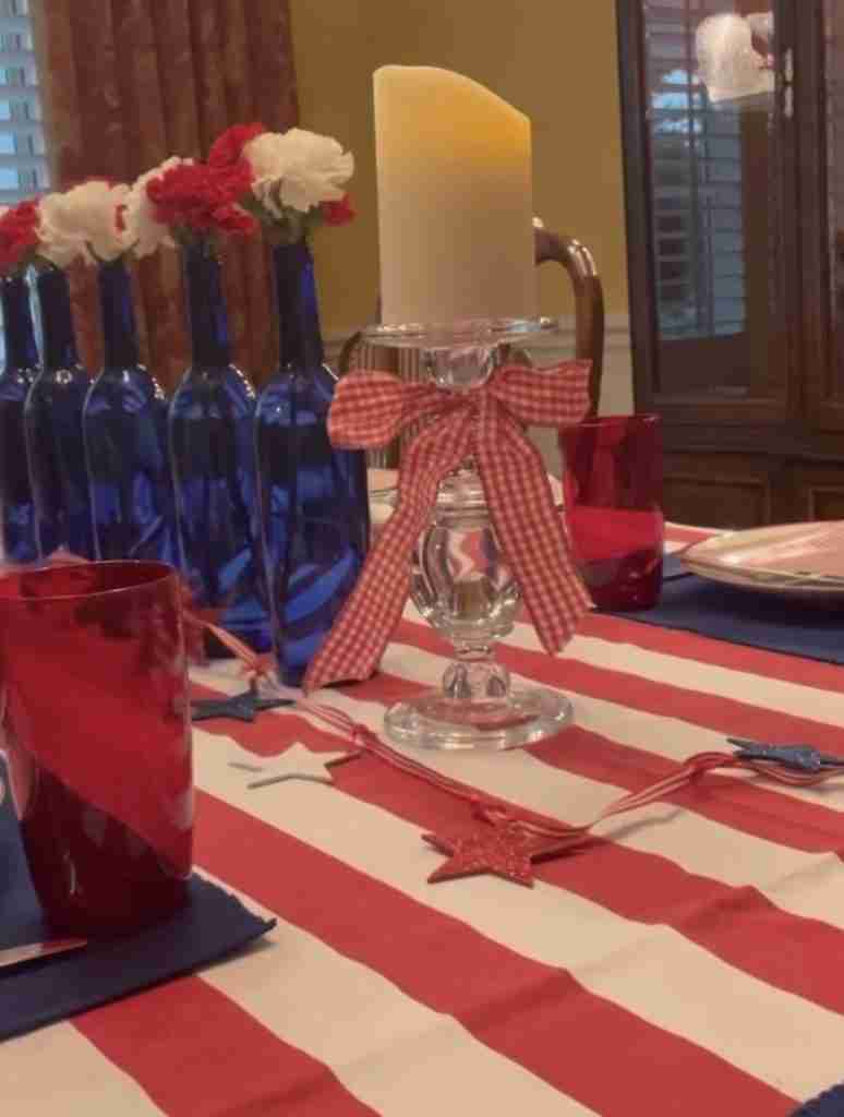 Here's a gorgeous 4th of July tablescape with a red striped tablecloth, blue napkins, and flag dessert plates,  There are red, white, and blue garlands, as well as bandana napkins.