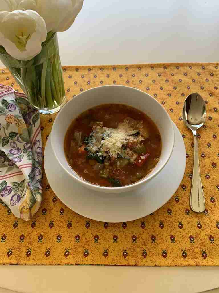 A  white ceramic bowl of ten vegetable soup.  The cowl is on a yellow print placemat, with a pretty print napkin and a vase filled with white tulips,