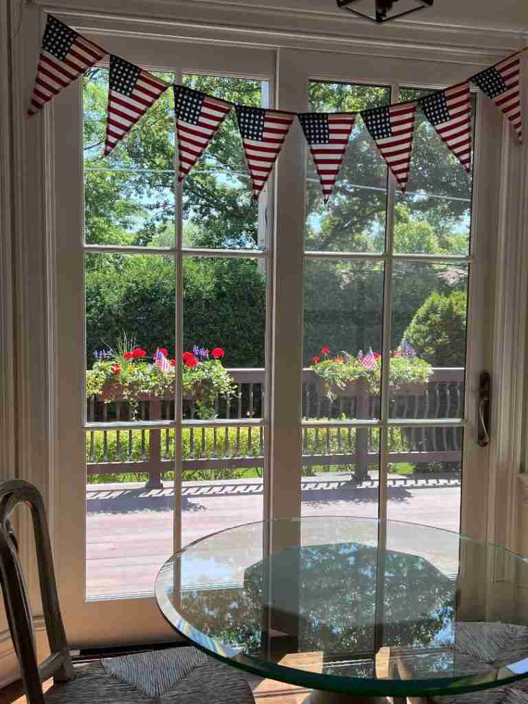This is a photo of my glass kitchen table for two overlooking my deck which has flower boxes is filled with red geraniums and blue spikey flowers.  Above the French doors are a patriotic Anerican flag pennant banner.